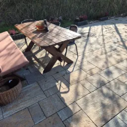 Ułożona kostka brukowa na tarasie z drewnianym stołem, ławką i dekoracjami ogrodowymi, w tle widoczna zieleń.