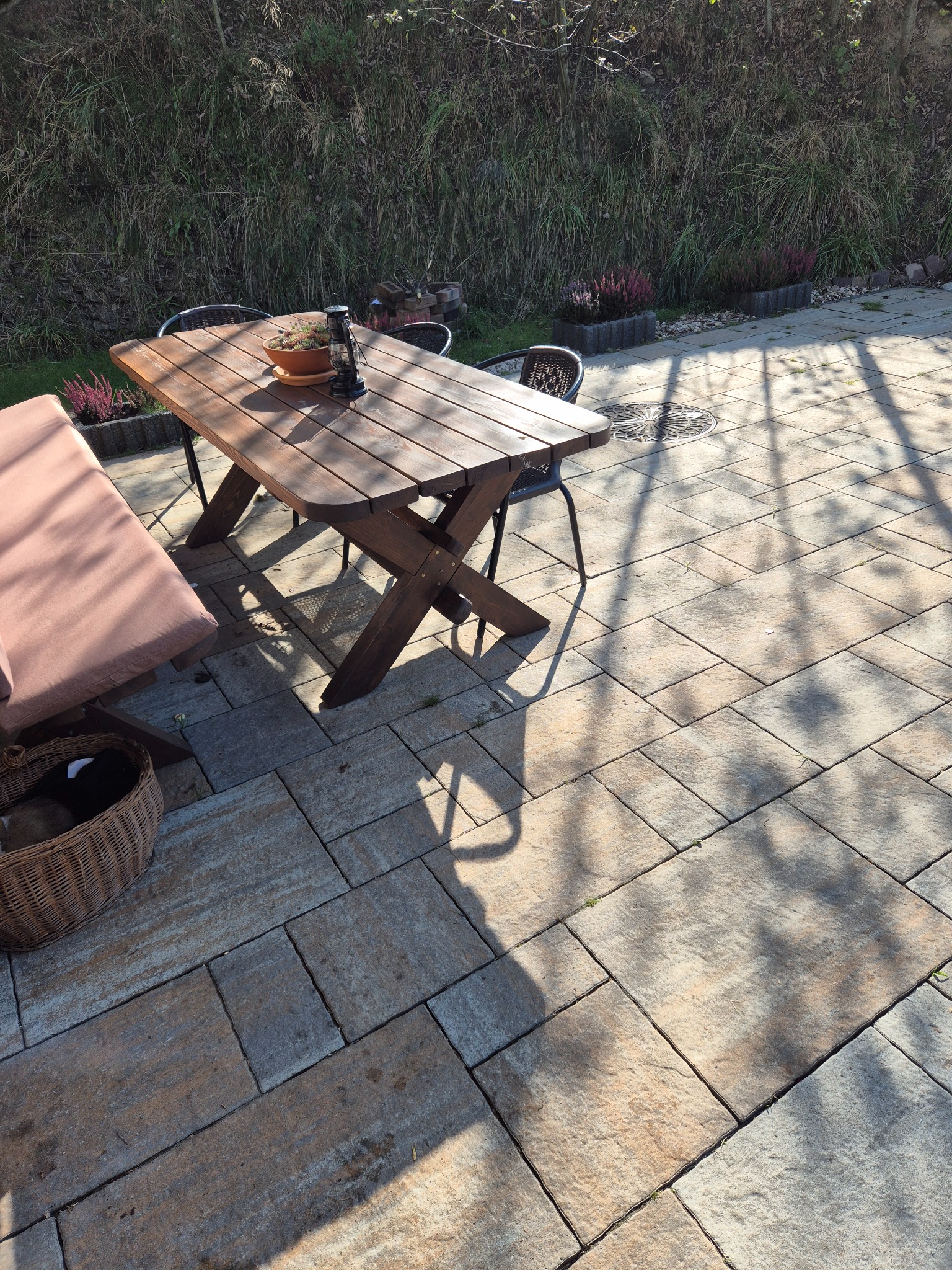 Ułożona kostka brukowa na tarasie z drewnianym stołem, ławką i dekoracjami ogrodowymi, w tle widoczna zieleń.