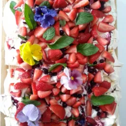 Kwadratowa Pavlova na drewnianej desce, udekorowana świeżymi truskawkami, borówkami, miętą i jadalnymi bratkami w różnych kolorach.