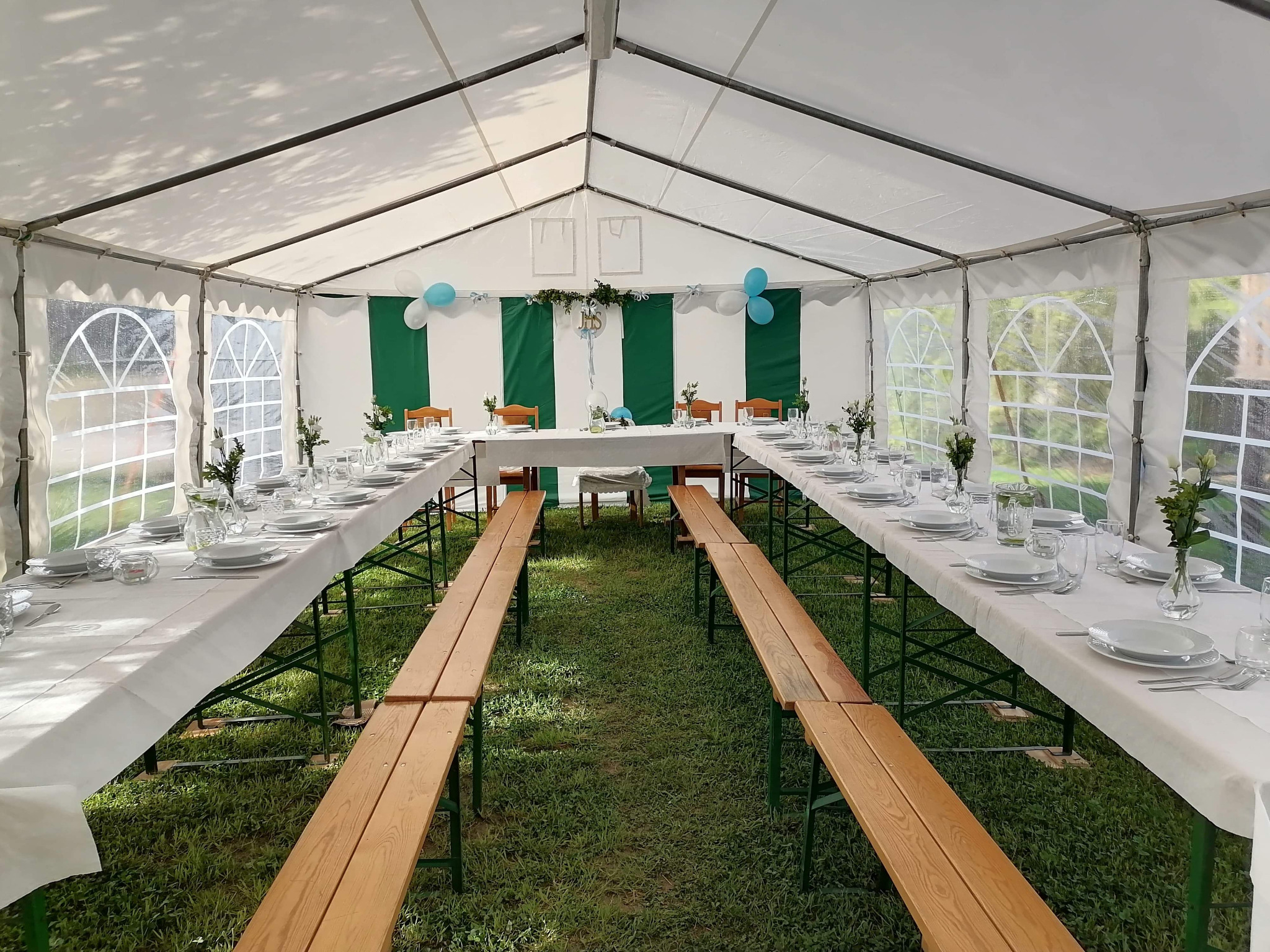 Ustawiony stół w białym namiocie z dekoracjami w odcieniach zieleni i błękitu, przygotowany na uroczystość na trawie.