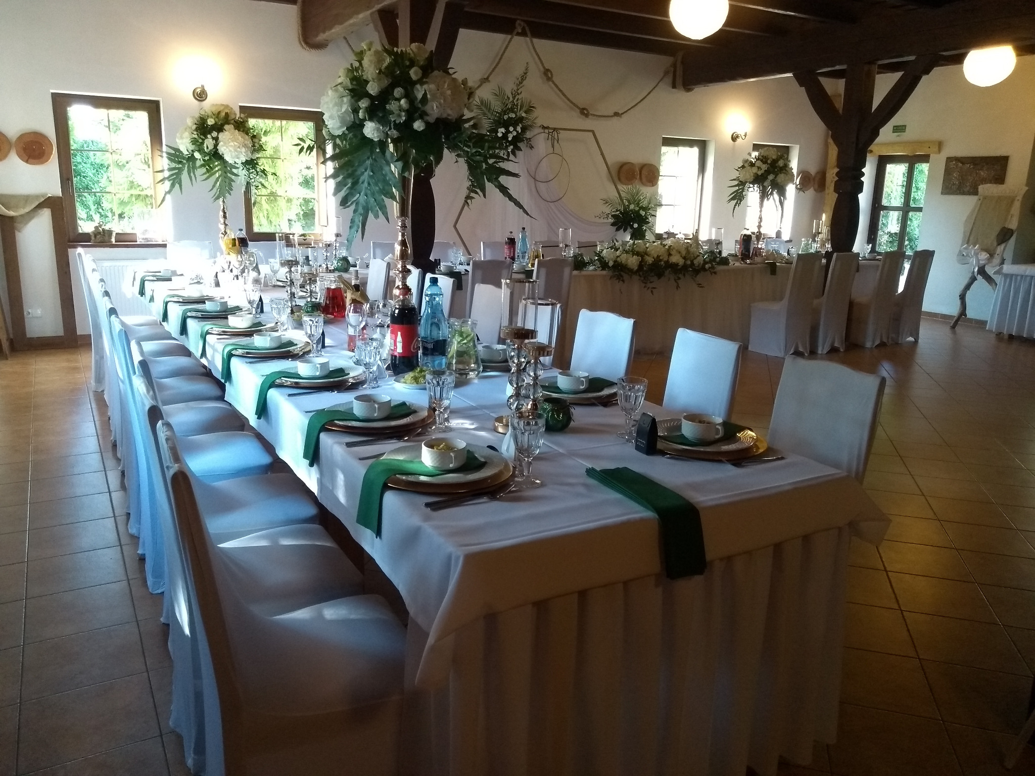 Uroczysta sala bankietowa z długim stołem nakrytym białym obrusem, zielonymi akcentami dekoracyjnymi, elegancką zastawą, kwiatowymi kompozycjami w wysokich wazonach i dekoracją ślubną w tle.