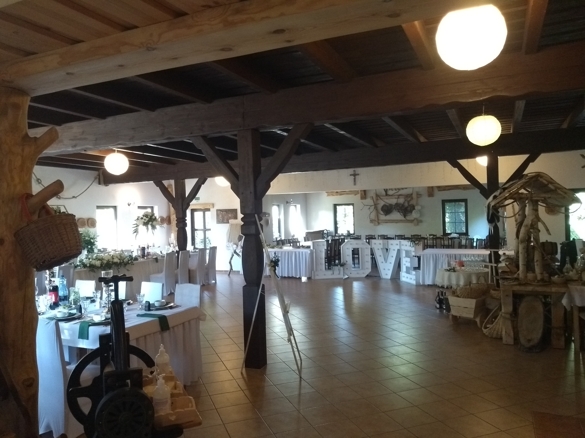 Sala bankietowa z dekoracjami ślubnymi: stoły z białymi obrusami, kwiatowe kompozycje, drewniane elementy dekoracyjne i podświetlane litery 'LOVE'.