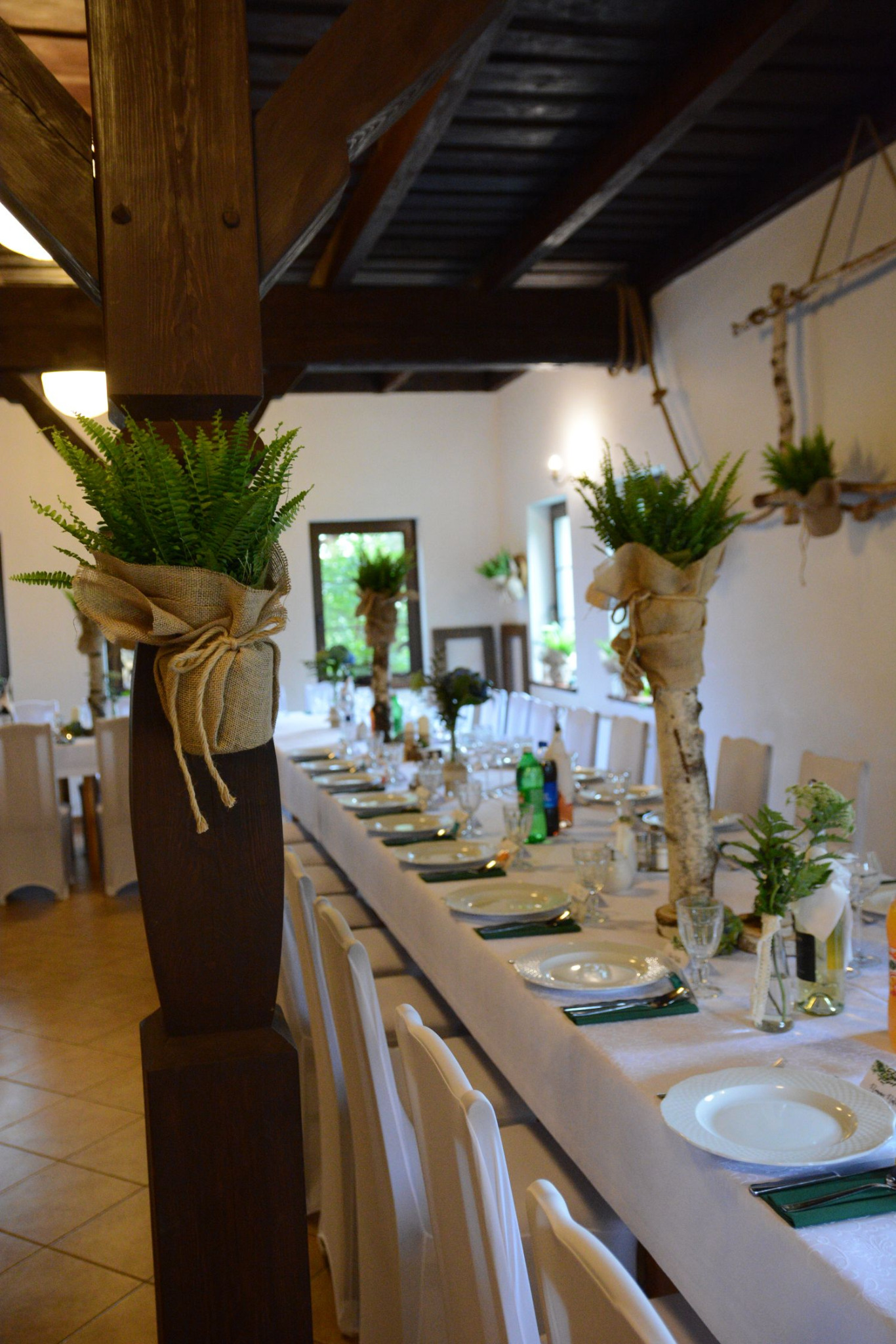 Długi stół nakryty białym obrusem, z elegancką zastawą i dekoracjami roślinnymi, w rustykalnym wnętrzu z drewnianymi elementami konstrukcyjnymi.