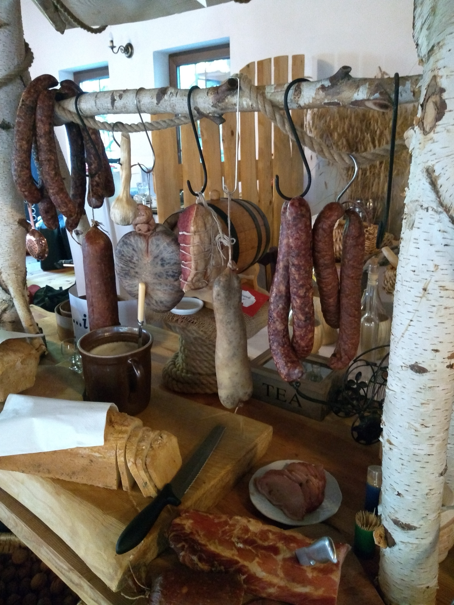 Rustykalna ekspozycja wędlin i pieczywa: wiszące kiełbasy na gałęzi brzozy, chleb na desce obok noża, plastry wędliny na talerzyku, całość w ciepłej tonacji.