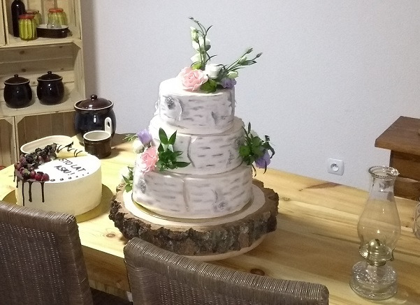 Trzypiętrowy tort weselny w stylu rustykalnym, imitujący korę brzozy, udekorowany kwiatami i umieszczony na drewnianym plastrze, obok tort z owocami w Brusach. W tle widoczny regał z naczyniami.