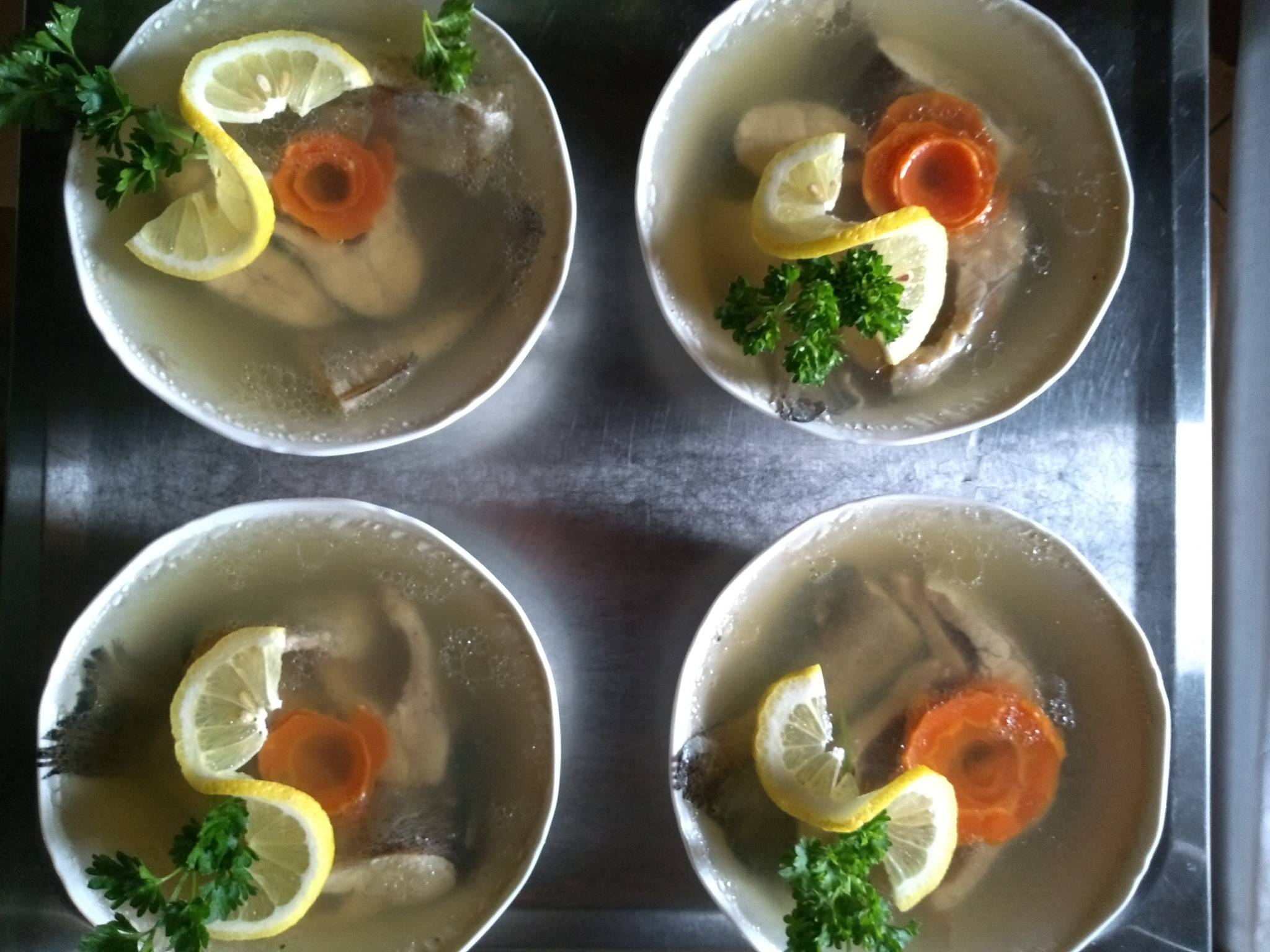 Cztery talerze galarety rybnej z dekoracją z plasterków cytryny w kształcie litery S, marchewki uformowanej w różyczkę i gałązki zielonej pietruszki, ułożone na metalowej tacy.