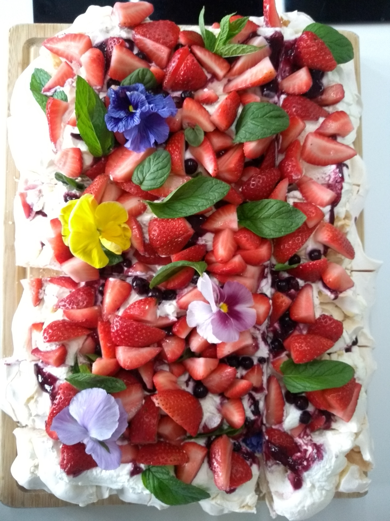 Kwadratowa Pavlova na drewnianej desce, udekorowana świeżymi truskawkami, borówkami, miętą i jadalnymi bratkami w różnych kolorach.