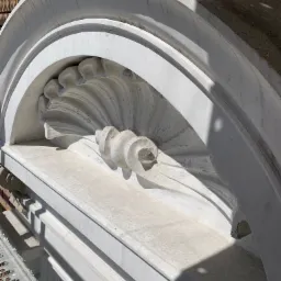Szczegółowe ujęcie odrestaurowanego, ozdobnego elementu elewacji w kształcie muszli, wykonanego z jasnego kamienia lub betonu, z widocznymi śladami cieni i fragmentem rusztowania.