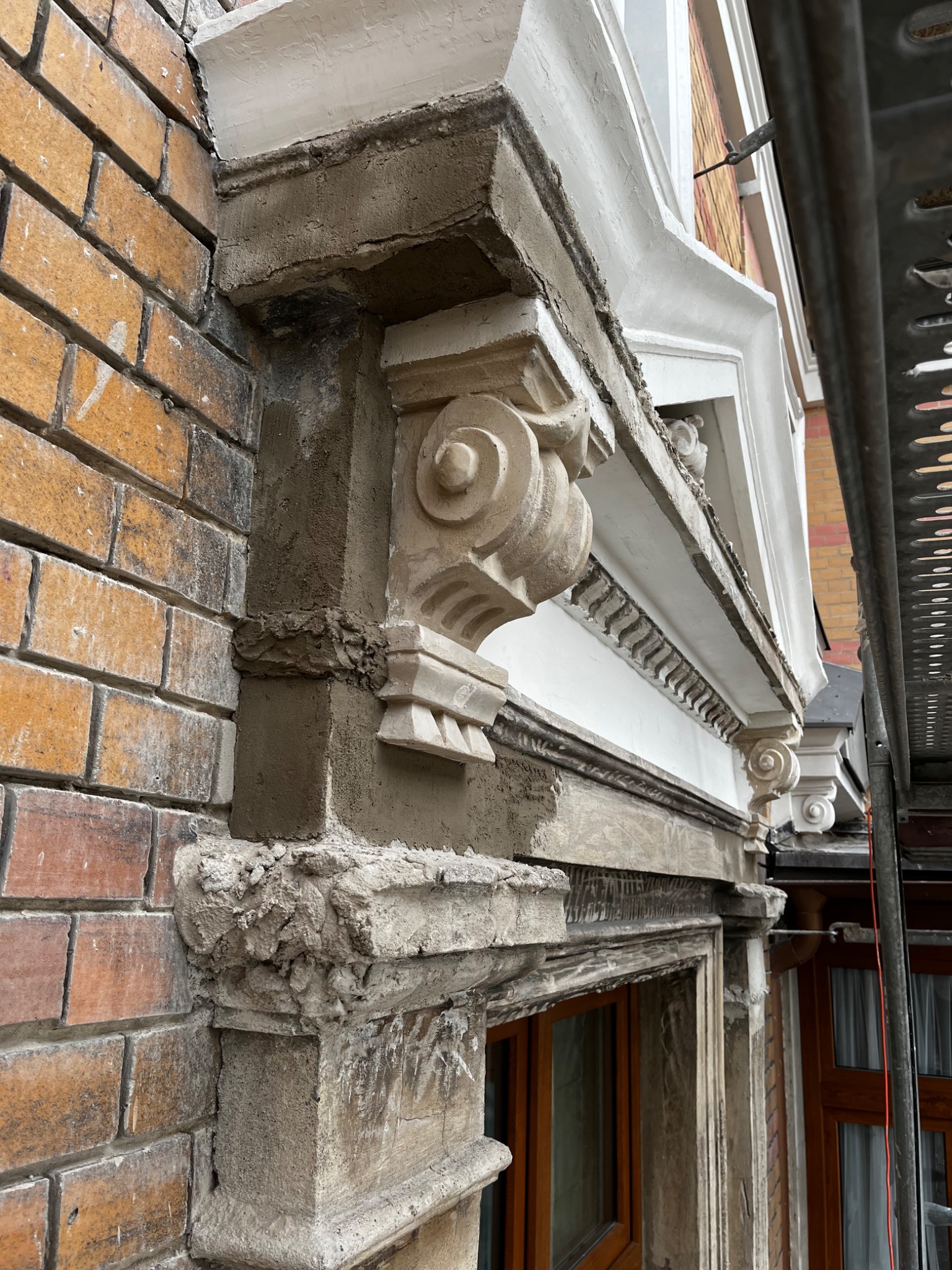 Renowacja detali architektonicznych elewacji: fragment gzymsu z dekoracyjnym wspornikiem w trakcie naprawy, widoczne ślady świeżego cementu i rusztowanie.