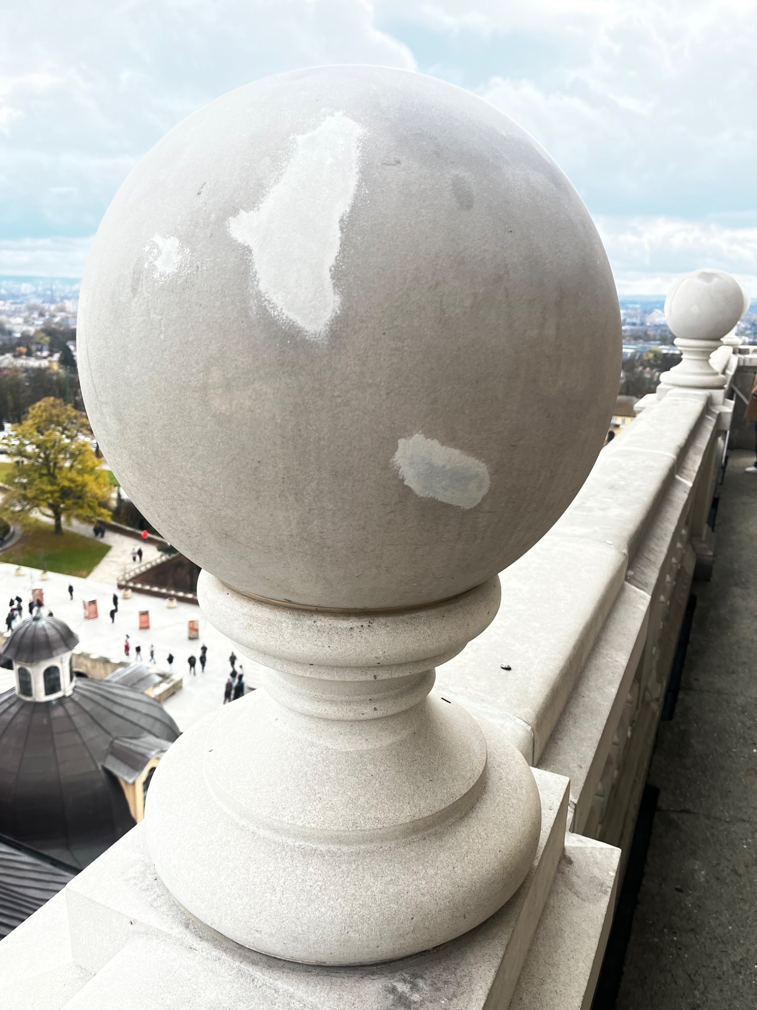 Kamienna balustrada z kulistymi zwieńczeniami, z widocznymi śladami napraw i uzupełnień, na tle miejskiego krajobrazu i kopuły budynku.