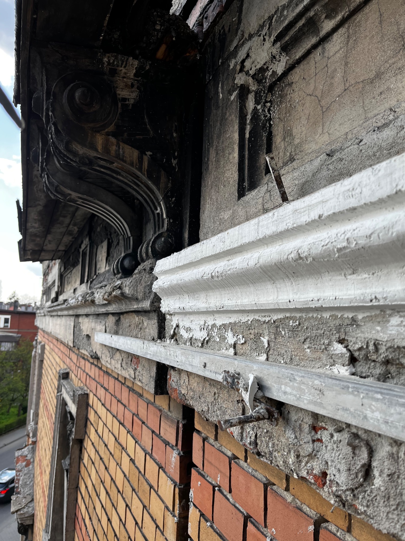 Fragment elewacji budynku z cegły i kamienia, ukazujący detale architektoniczne w trakcie prac, z widocznymi elementami dekoracyjnymi i śladami uszkodzeń oraz nowymi elementami mocującymi.