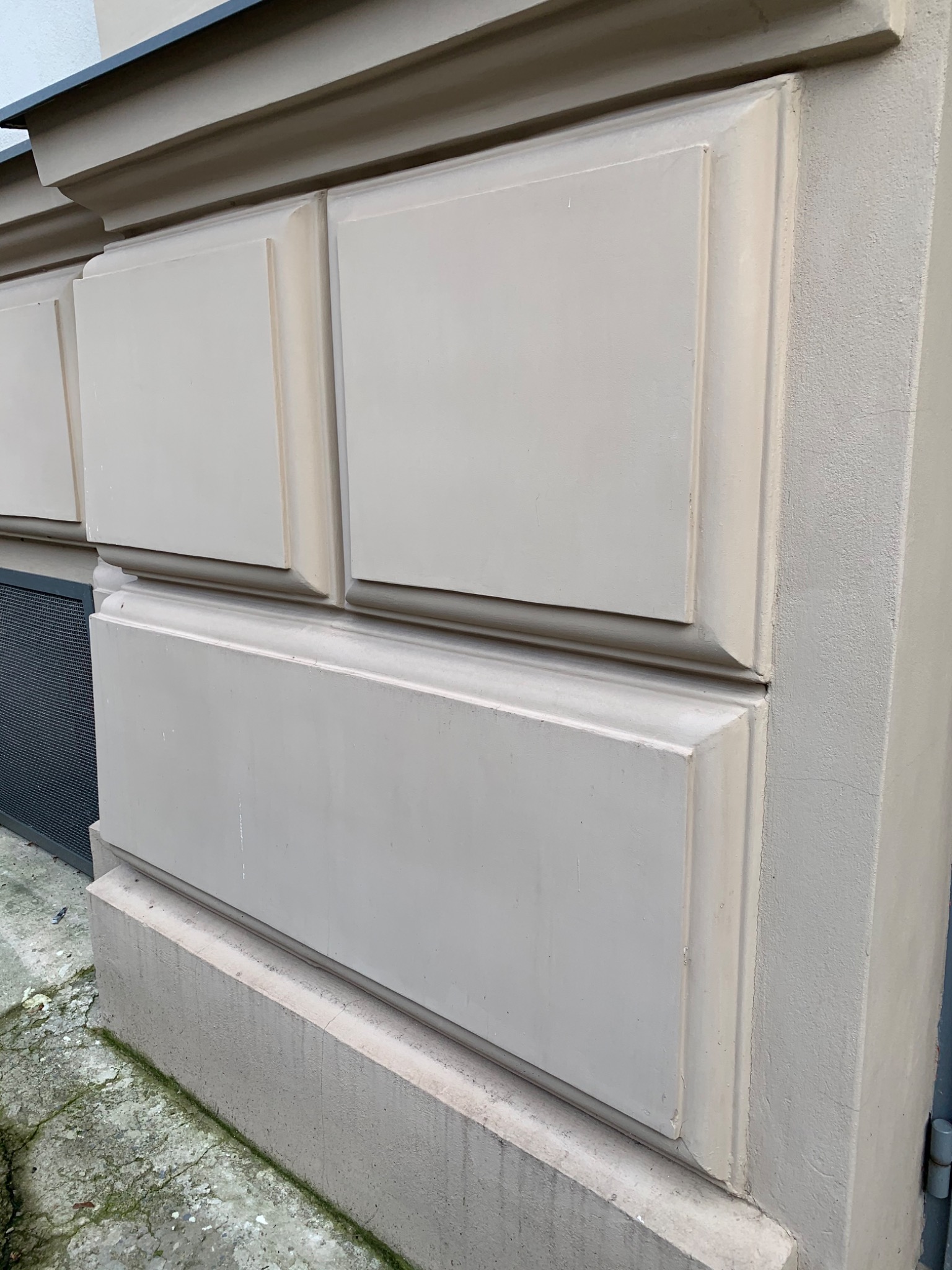 Fragment elewacji budynku z dekoracyjnymi panelami w odcieniach beżu, widoczne zabrudzenia na cokole i chodniku.