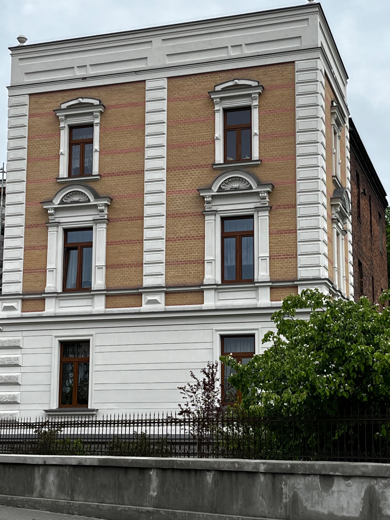 Odnowiona elewacja kamienicy z cegły klinkierowej z białymi elementami dekoracyjnymi, widoczna z poziomu ulicy, z fragmentem ogrodzenia i zieleni w kadrze.