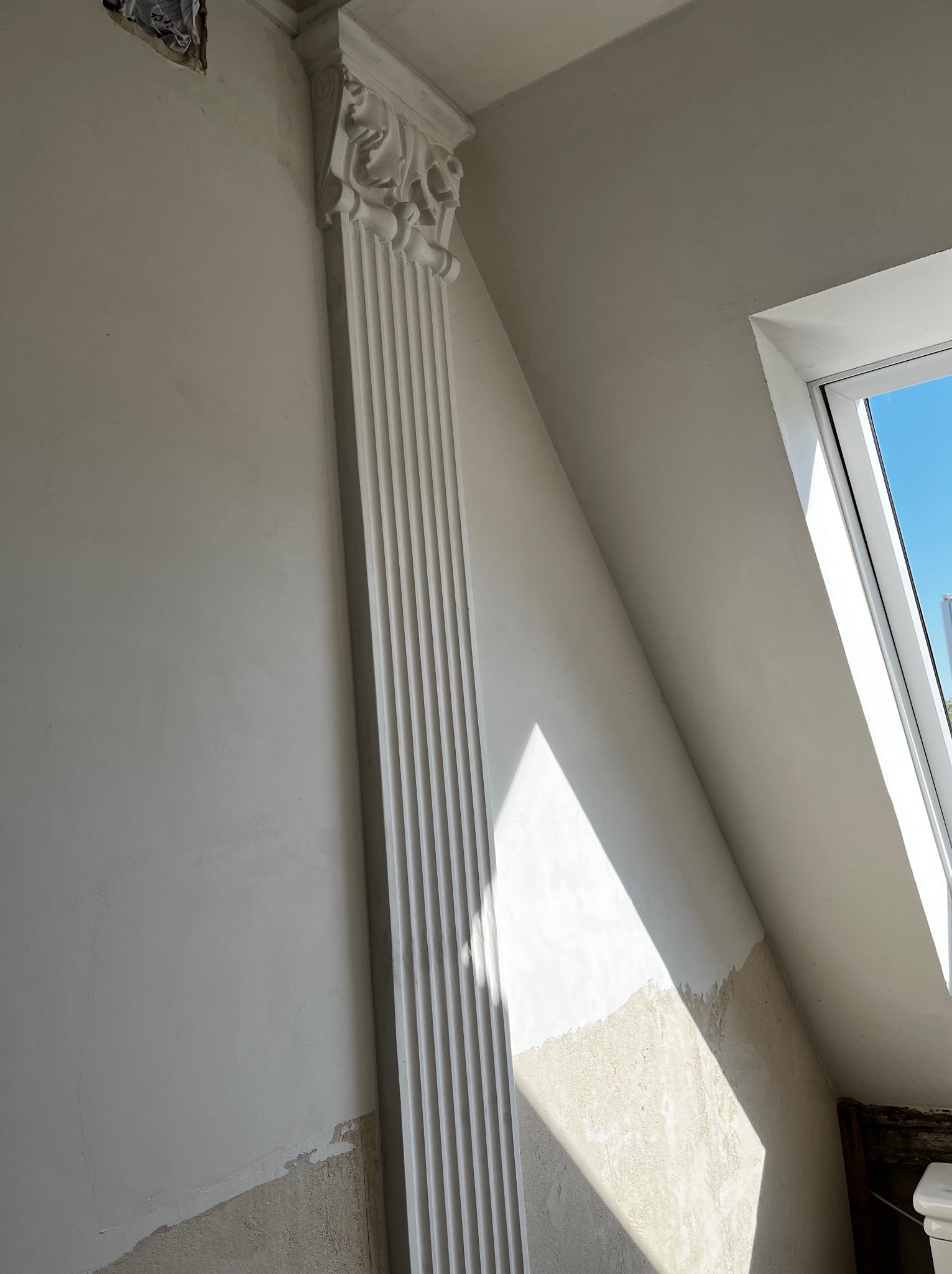 Wnętrze poddasza w trakcie prac wykończeniowych, widoczna kolumna z dekoracyjnym kapitelem, ściana częściowo otynkowana, fragment okna dachowego z widokiem na błękitne niebo.