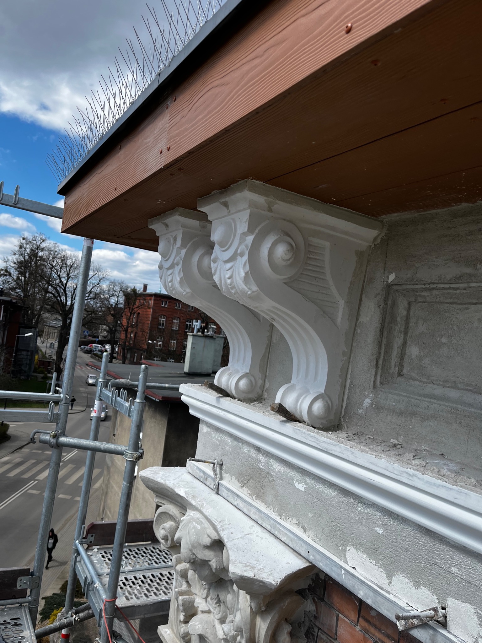 Odnowiona elewacja budynku z dekoracyjnymi wspornikami i zabezpieczeniem przed ptakami, widok z rusztowania na ulicę.