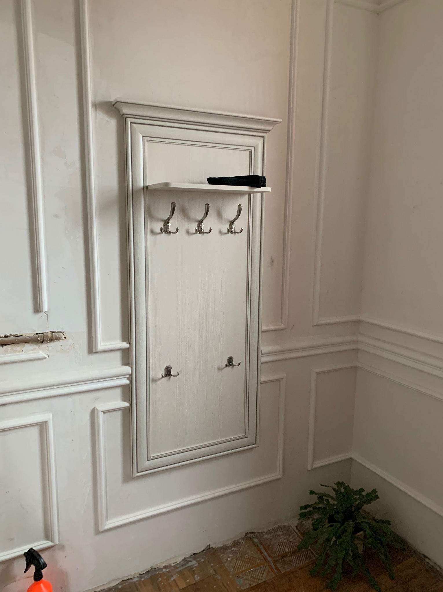 Biała, klasyczna szafka z wieszakami i półką, zamontowana na ścianie z dekoracyjnymi listwami, w trakcie renowacji. Widoczne ślady prac na ścianie i podłodze, oraz roślina w doniczce w rogu pokoju.