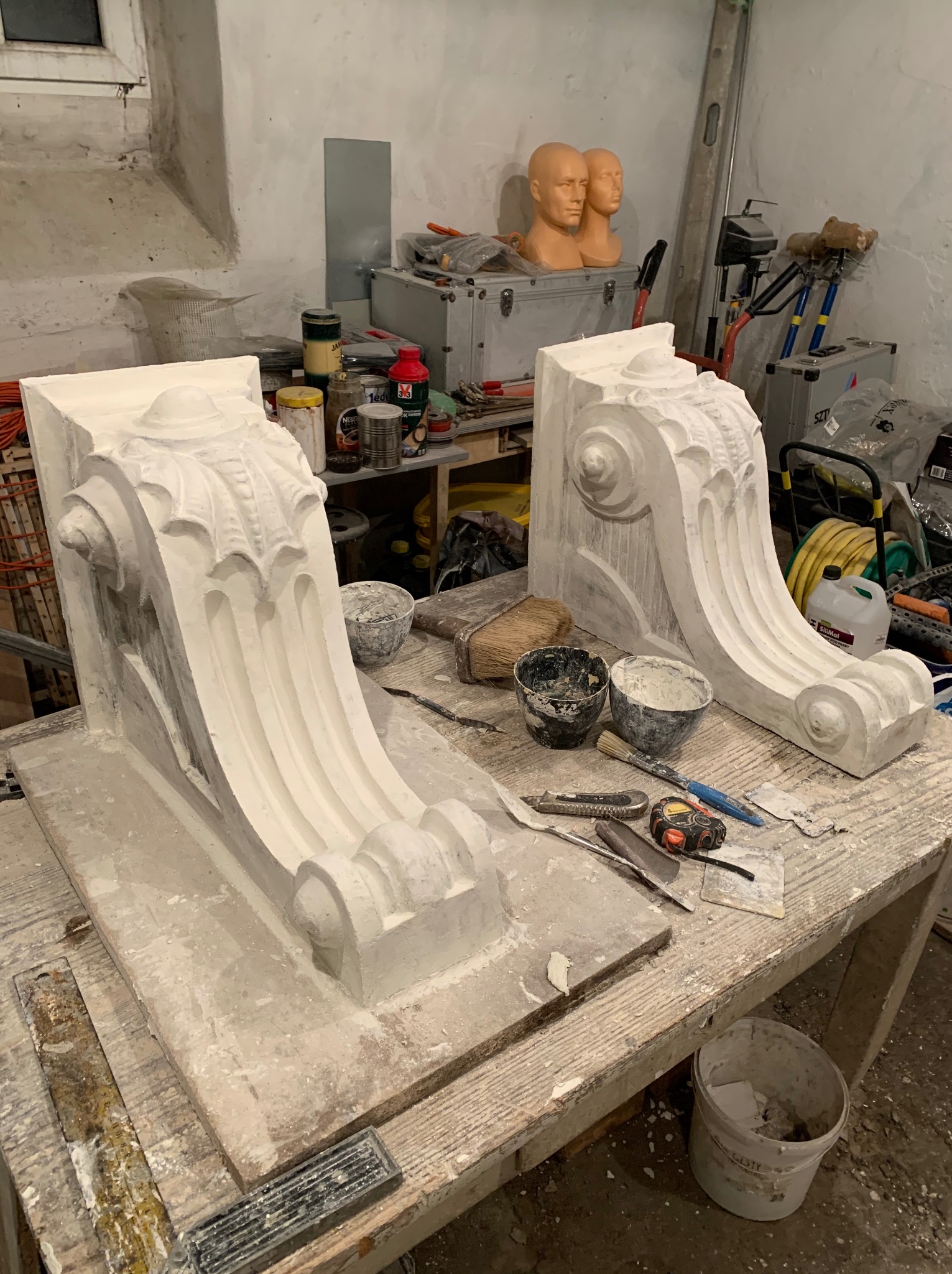 Dwa gipsowe wsporniki z ornamentami leżące na stole w warsztacie, otoczone narzędziami i materiałami budowlanymi, w tle widoczne manekiny głów.