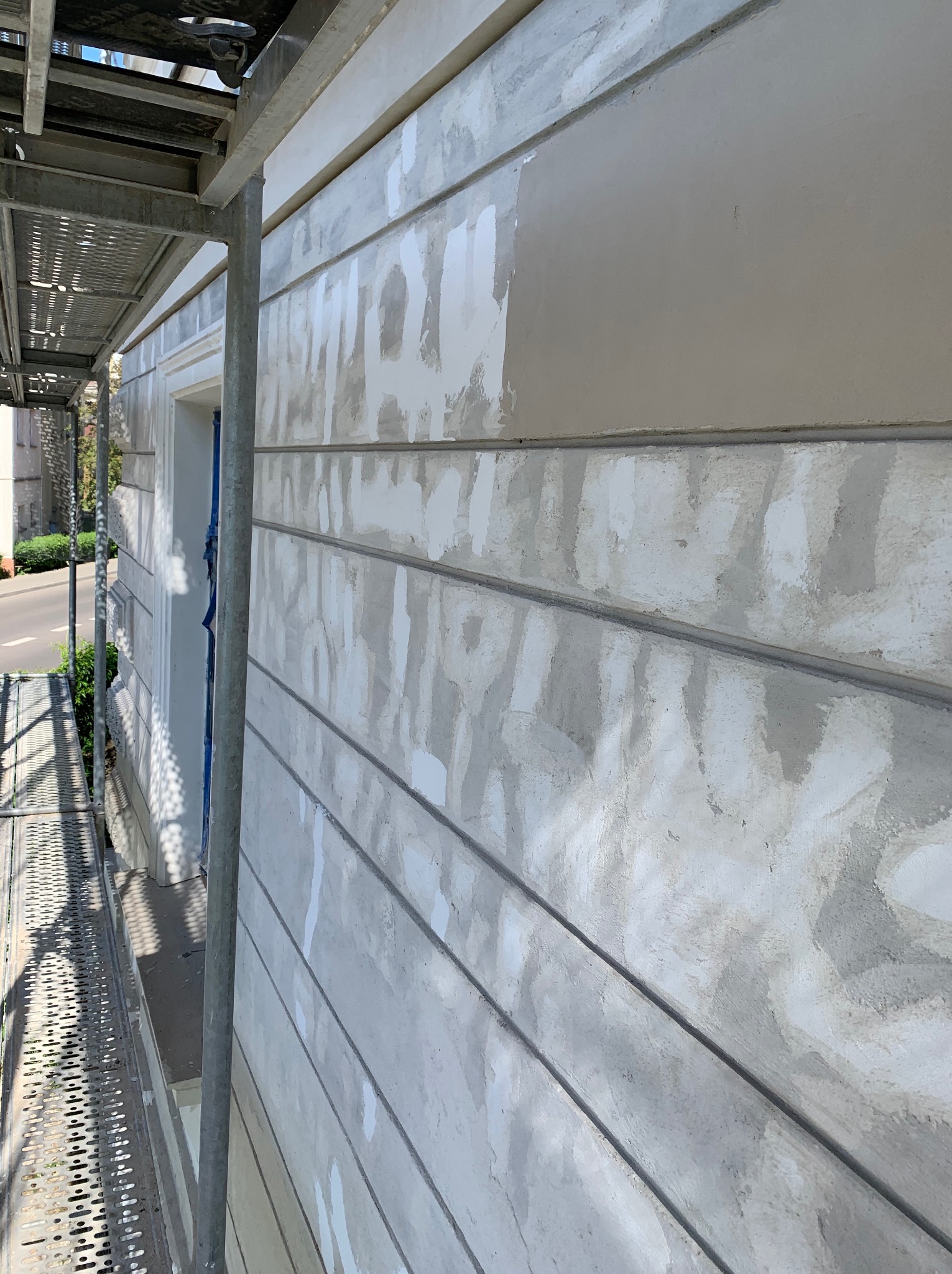 Fragment elewacji budynku z widocznymi śladami prac przygotowawczych do malowania, częściowo pokrytej nową warstwą farby, z metalowym rusztowaniem po lewej stronie.