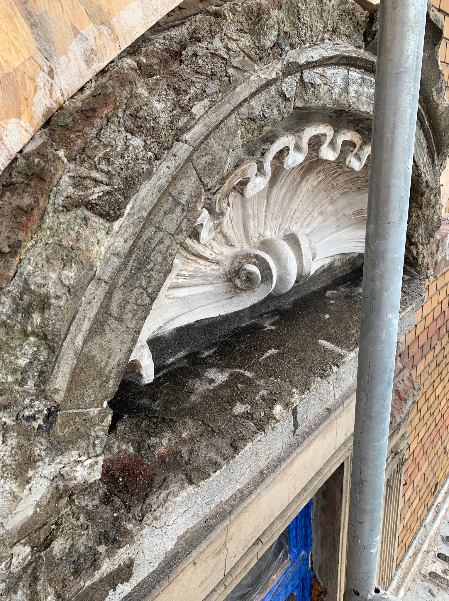 Fragment elewacji budynku z dekoracyjnym elementem w kształcie muszli, w trakcie prac remontowych. Widoczne uszkodzenia, surowe materiały i rusztowanie.