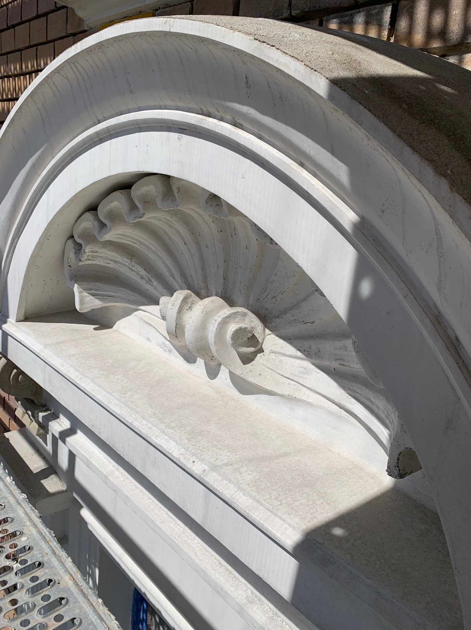 Szczegółowe ujęcie odrestaurowanego, ozdobnego elementu elewacji w kształcie muszli, wykonanego z jasnego kamienia lub betonu, z widocznymi śladami cieni i fragmentem rusztowania.