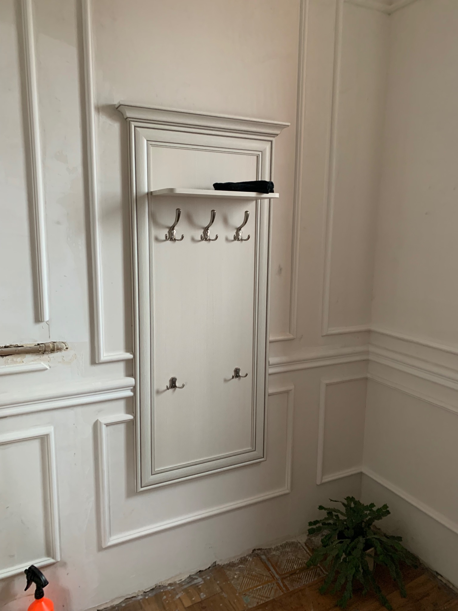 Biały panel ścienny z wieszakami, zamontowany na ścianie z dekoracyjnymi listwami. Widoczne ślady prac remontowych i doniczka z rośliną w rogu.