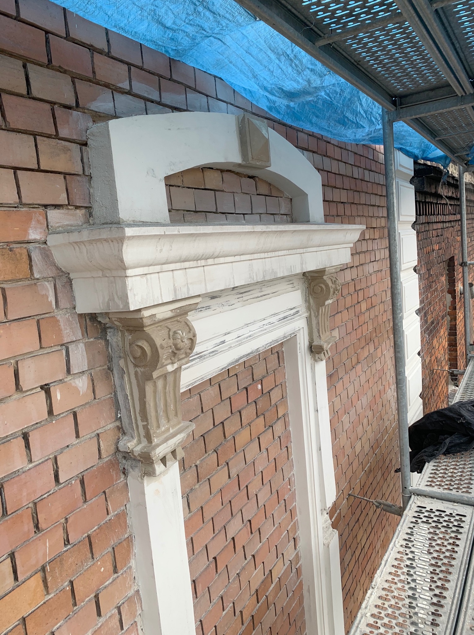 Fragment odnawianej elewacji z cegły z dekoracyjnym elementem architektonicznym nad oknem, widoczne rusztowanie i plandeka ochronna.