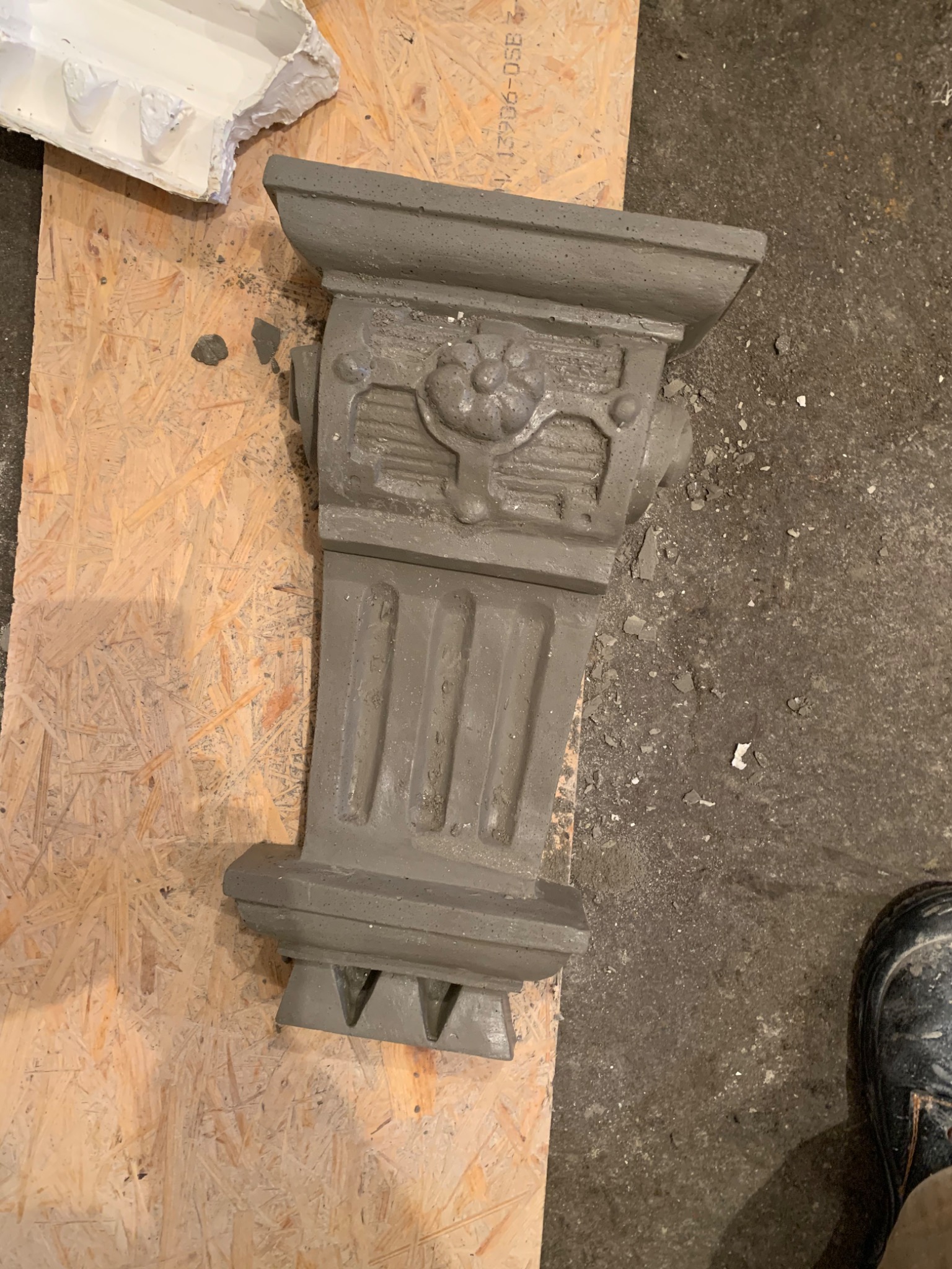 Szary, betonowy element dekoracyjny elewacji w stylu klasycznym, leżący na płycie OSB, obok widoczny fragment formy odlewniczej i fragment buta roboczego.