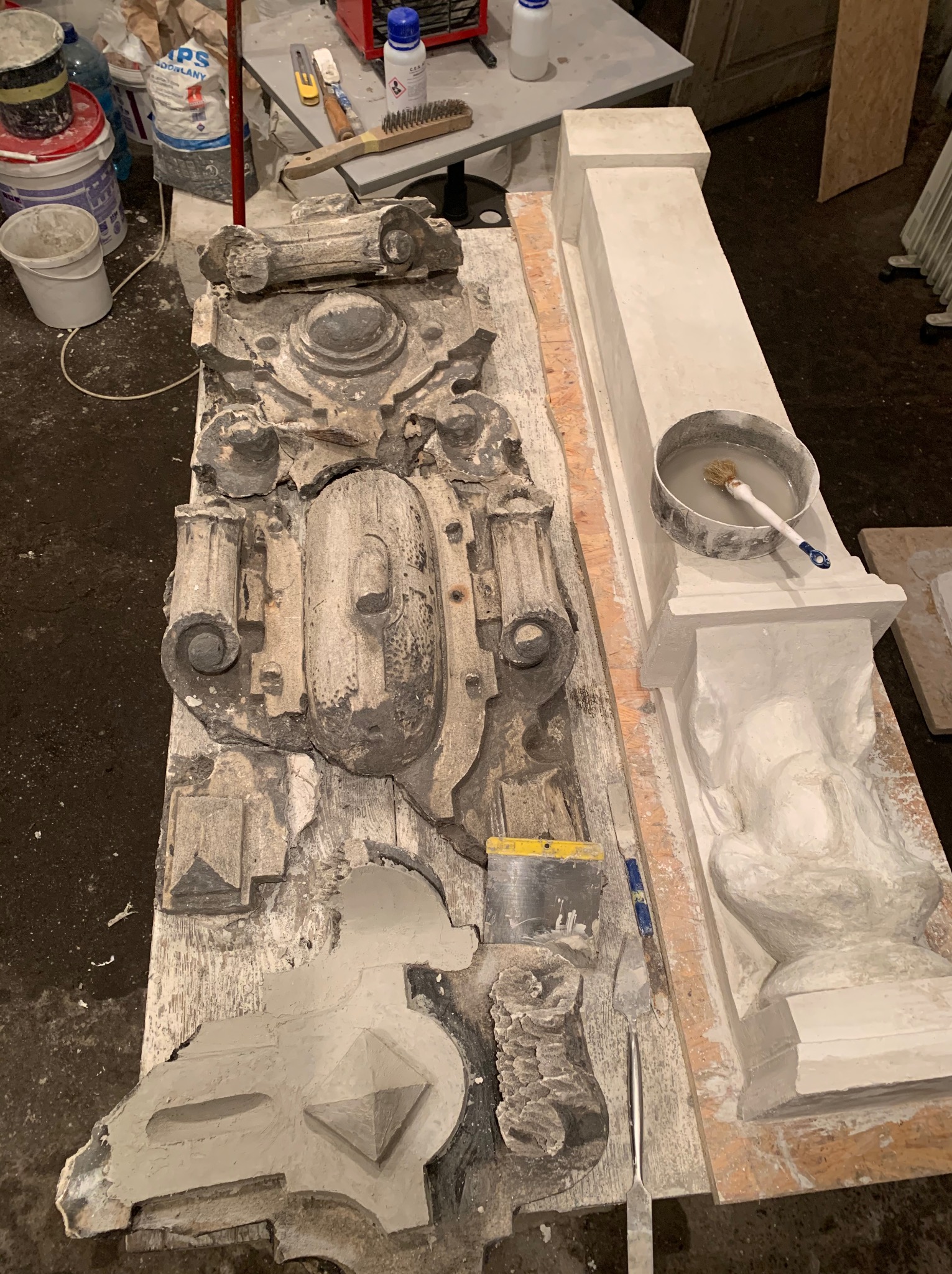 Fragmenty architektoniczne, w tym kolumna z kapitelem i ozdobny element elewacji, w trakcie czyszczenia i naprawy. Widoczne narzędzia i materiały, takie jak pędzle, szpachelki i zaprawy.