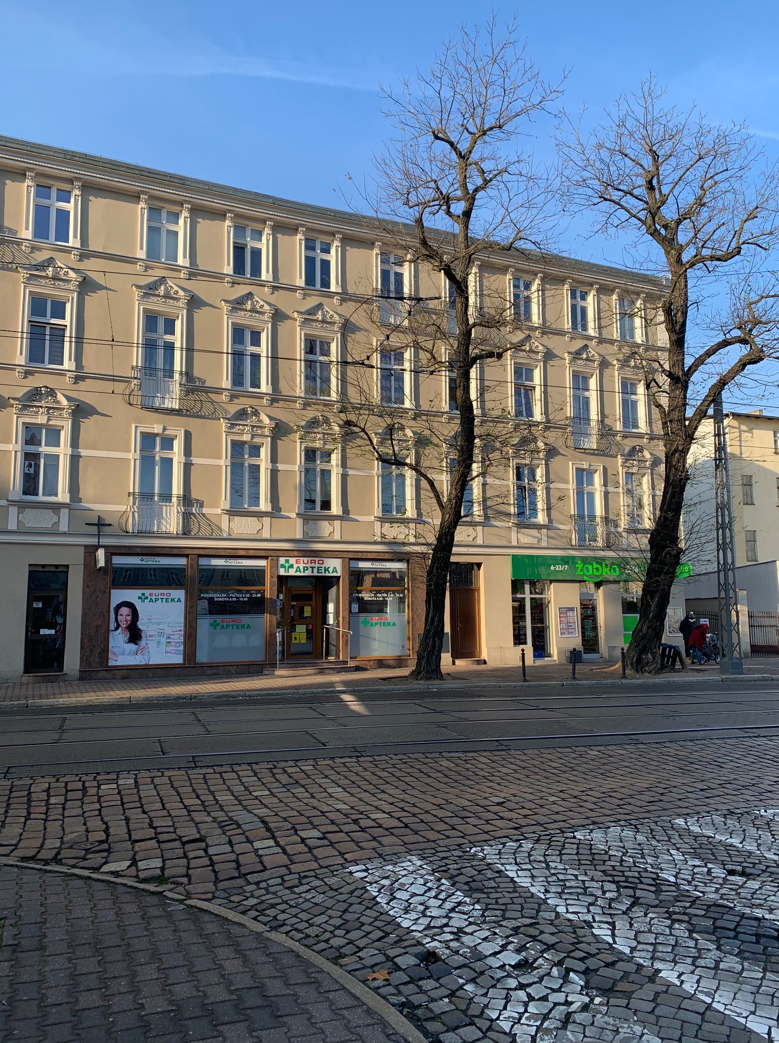 Odnowiona kamienica z beżową elewacją, ozdobnymi detalami okiennymi i balkonami, widoczna z perspektywy ulicy z brukowanym chodnikiem i torami tramwajowymi.