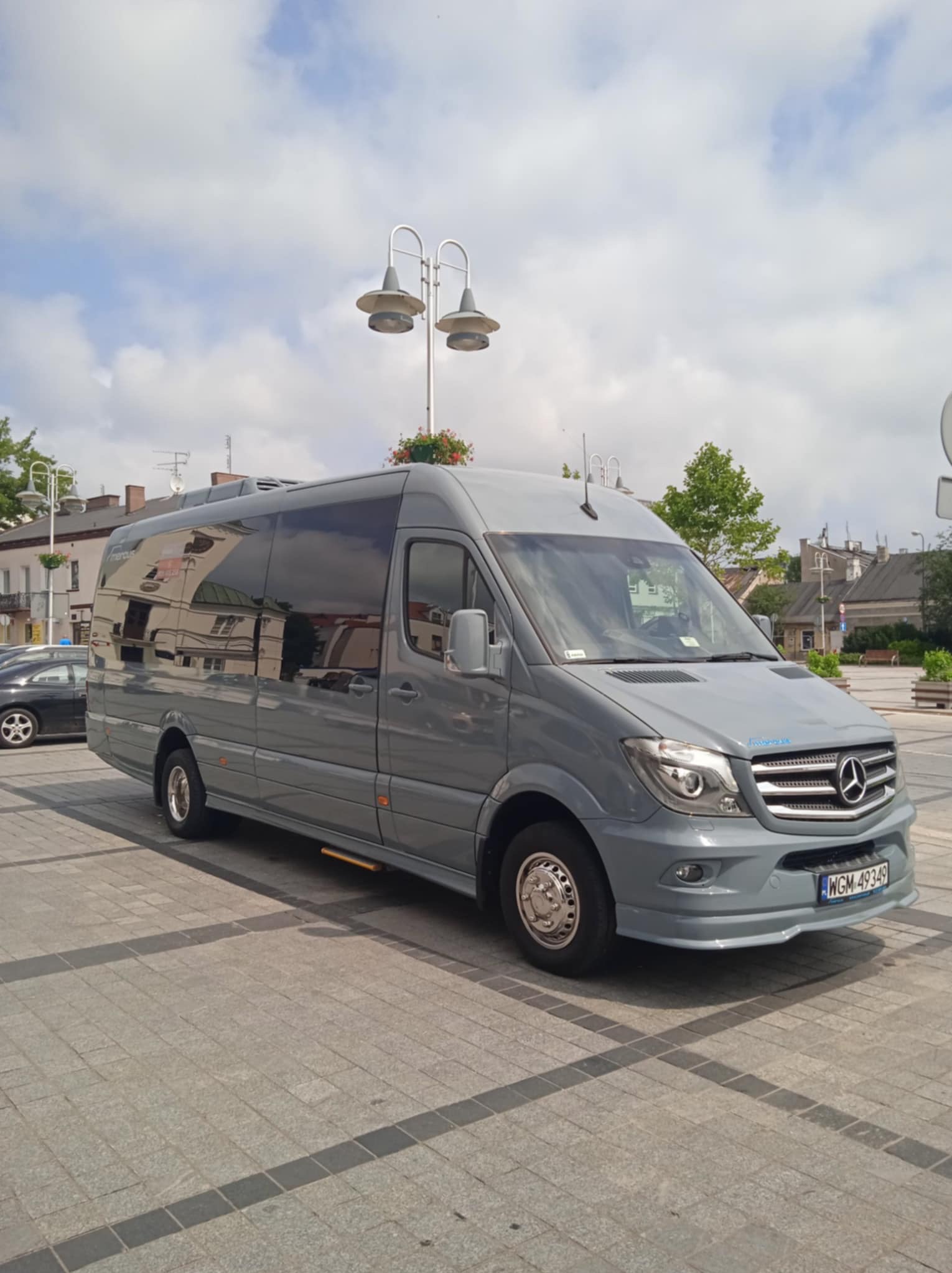 Szary bus Mercedes-Benz Sprinter z przyciemnianymi szybami, zaparkowany na placu miejskim z widocznym budynkiem w tle. Tablica rejestracyjna WGM.