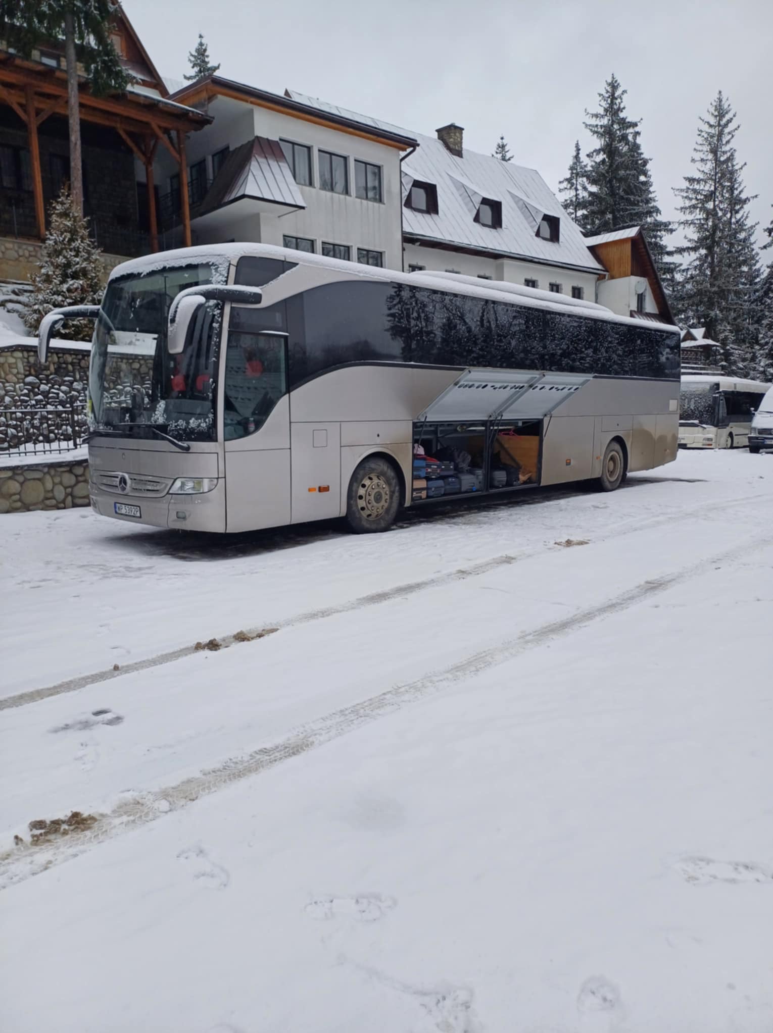 Srebrny autokar Mercedes-Benz z otwartym bagażnikiem na śnieżnym parkingu, w tle budynek w stylu górskim i drzewa. Widoczne ślady opon na śniegu.