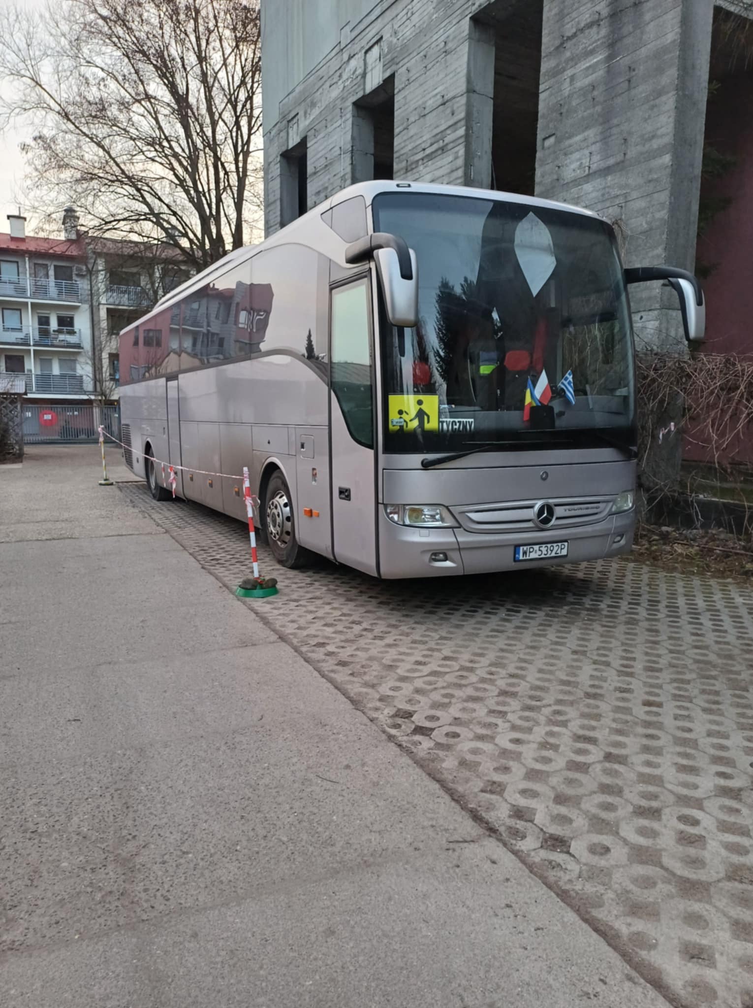 Srebrny autokar Mercedes-Benz z tabliczką 'Tyczyny' i flagami (Rumunia, Grecja) zaparkowany w Wiskitkach. Widoczny znak ostrzegawczy dla dzieci.