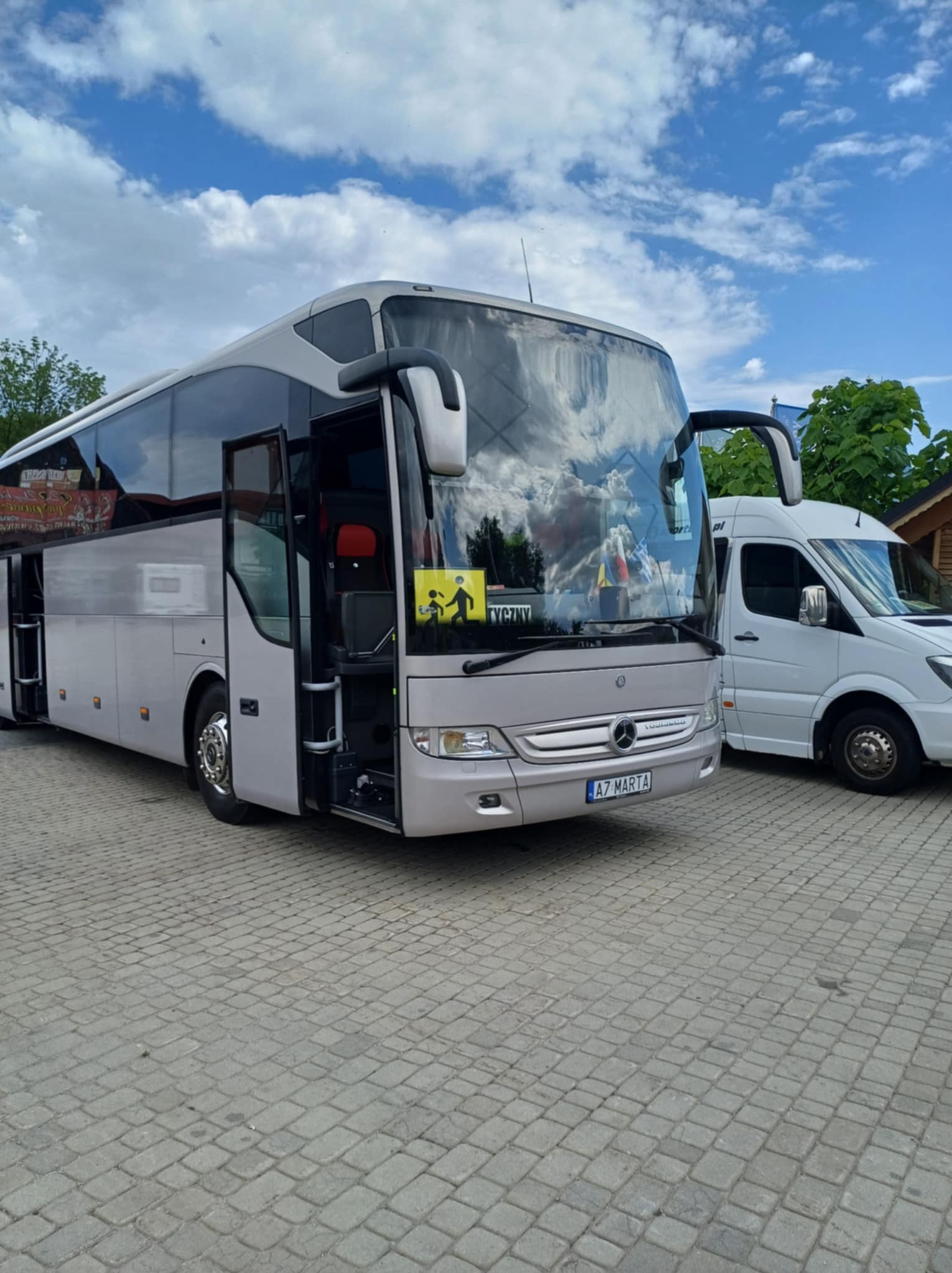 Srebrny autokar Mercedes-Benz z otwartymi drzwiami, zaparkowany obok białego busa na brukowanym placu w słoneczny dzień. Widoczna tablica rejestracyjna.