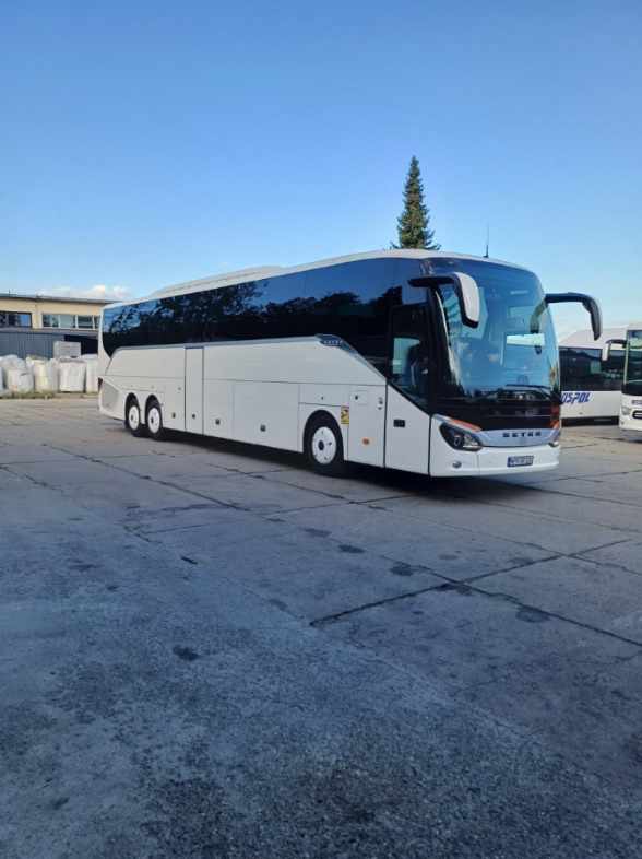Nowoczesny, biały autokar Setra na betonowym placu, z widocznym fragmentem innego autobusu w tle i budynkiem. Błękitne niebo i drzewo iglaste w tle.