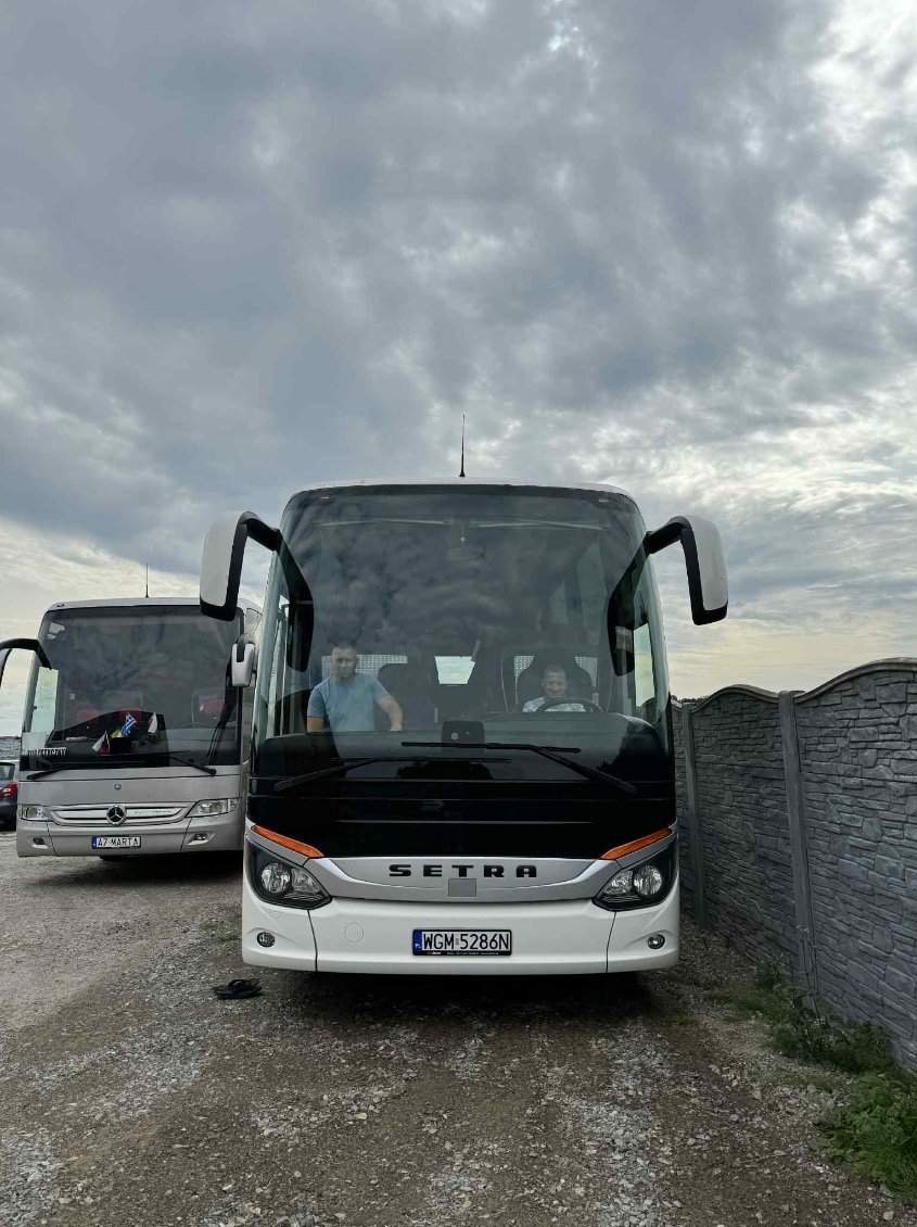 Dwa autokary Setra na żwirowym parkingu, z widocznymi kierowcami w kabinach. Jeden biały z czarnym pasem, drugi srebrny. W tle szary, pochmurny dzień.