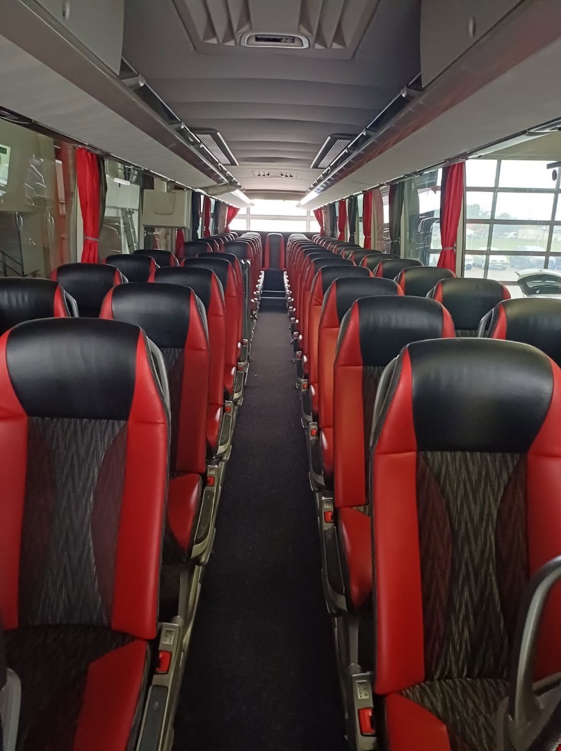 Wnętrze autobusu z rzędami czarno-czerwonych foteli, widok z przodu na tył pojazdu. Czerwone zasłonki przy oknach, czarna wykładzina podłogowa.
