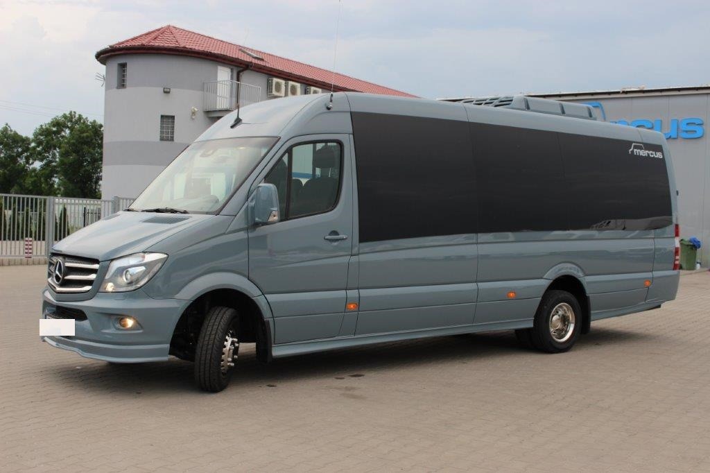 Szary bus Mercedes Sprinter z przyciemnianymi szybami, zaparkowany na kostce brukowej przed budynkiem z szyldem firmy.