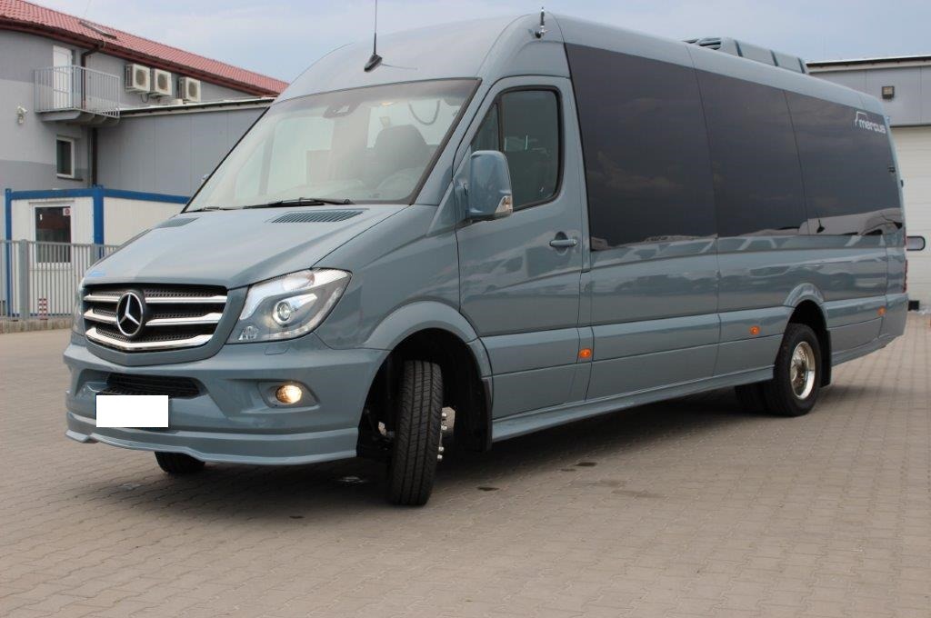 Szary Mercedes Sprinter z przyciemnianymi szybami, zaparkowany na kostce brukowej, z widocznym logo na masce i dodatkowym oświetleniem w dolnej części zderzaka.