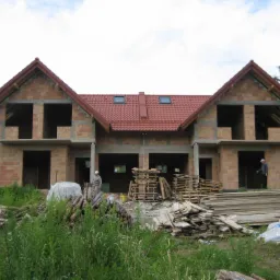 Surowy stan bliźniaczego domu z czerwonej cegły z dachem z czerwonej dachówki, widoczne drewniane palety i deski, dwóch pracowników wewnątrz budynku.