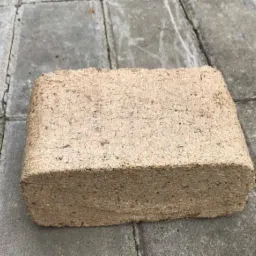 Pojedyncza kostka brykietu drzewnego, leżąca na szarym brukowanym podłożu, widoczna faktura sprasowanych trocin.