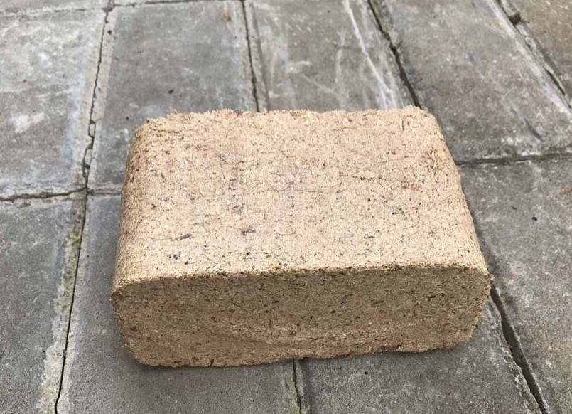 Pojedyncza kostka brykietu drzewnego, leżąca na szarym brukowanym podłożu, widoczna faktura sprasowanych trocin.