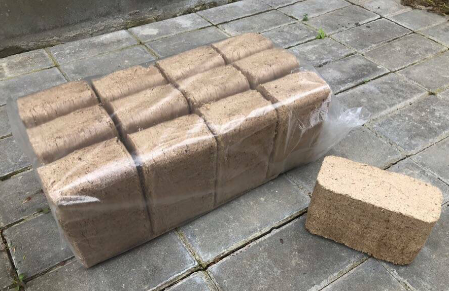 Pakiet sprasowanego materiału opałowego w folii, jeden element leży obok na szarym bruku.