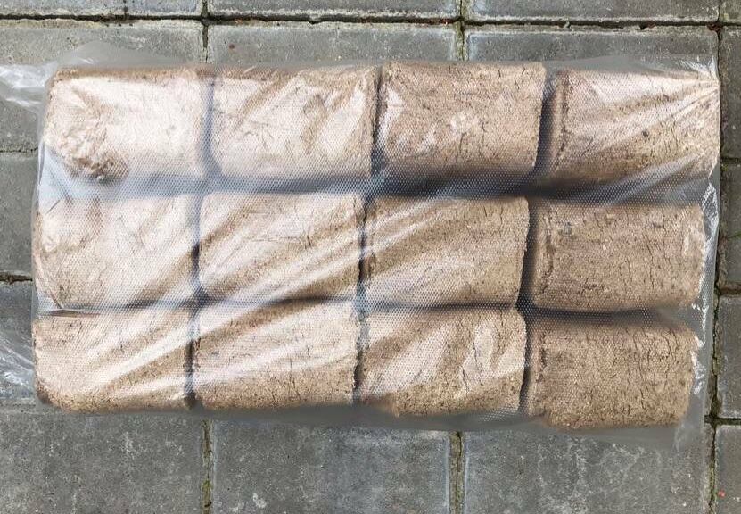 Pakiet dwunastu kostek prasowanego materiału opałowego z widoczną strukturą drewna, zapakowany w przezroczystą folię z perforacją, ułożony na szarej kostce brukowej.