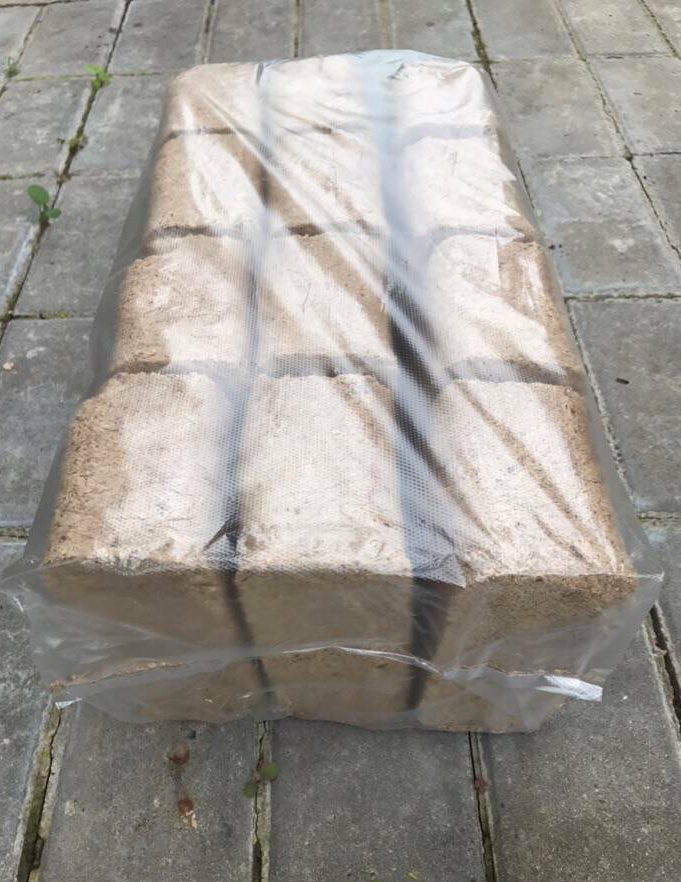 Paleta sprasowanego drewna w przezroczystej folii, ułożona na szarym bruku z widocznymi chwastami w spoinach.