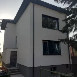 Dwukondygnacyjny dom z jasną elewacją i ciemną podmurówką, widok z zewnątrz, z widocznymi oknami z czarnymi ramami i elementami dachu w kolorze grafitowym, z fragmentem zieleni w tle.