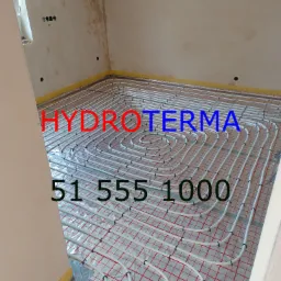 Białe rury ogrzewania podłogowego ułożone spiralnie na folii z czerwoną kratką, widoczne przyłącza z rurami w czerwonej i niebieskiej izolacji, ściany w trakcie wykończenia.