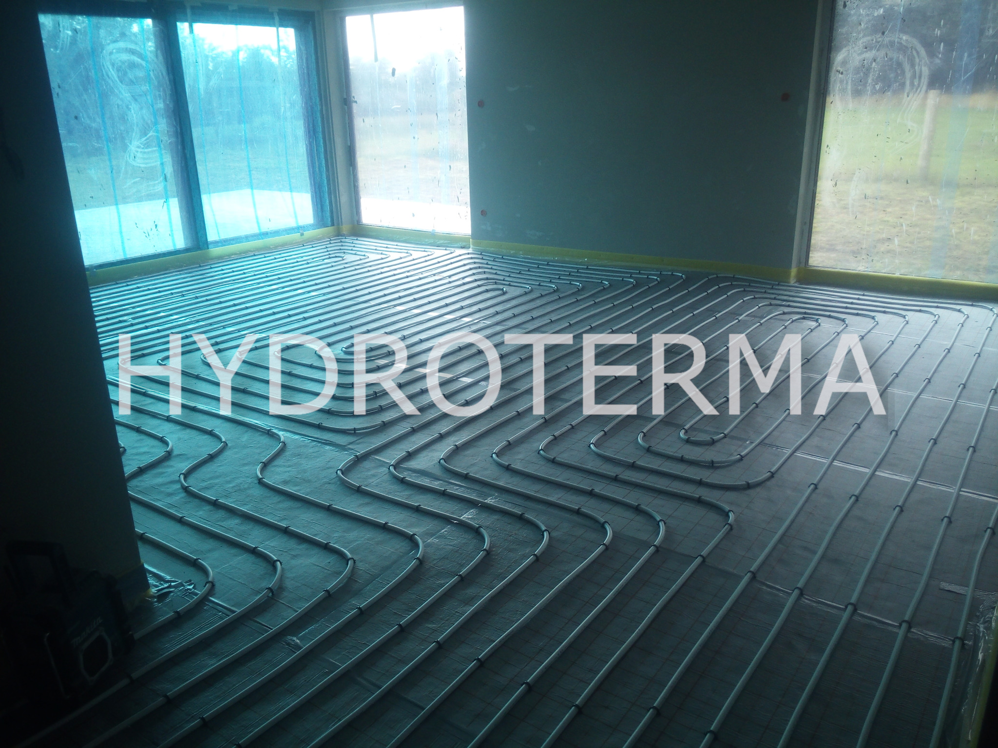 Równomiernie rozłożone, białe rurki ogrzewania podłogowego na czarnej macie izolacyjnej w surowym pomieszczeniu z oknami, zabezpieczonymi folią malarską, widoczne narzędzie Makita w lewym dolnym rogu.