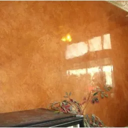 Ściana w odcieniach brązu z efektem dekoracyjnym, odbijająca światło okien, z rośliną doniczkową w rogu.