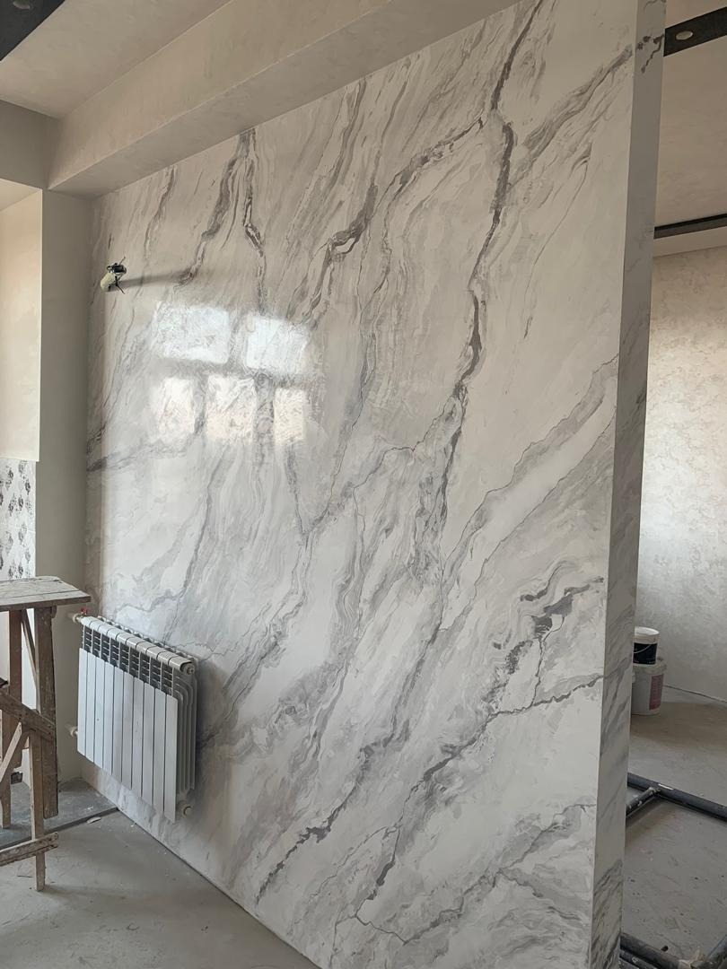 Ściana w pokoju pokryta dekoracyjnym tynkiem imitującym marmur, z widocznym grzejnikiem i elementami rusztowania, uchwycona z perspektywy lekko z dołu.