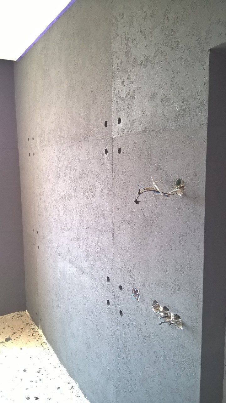 Ściana wewnętrzna wykończona szarą, dekoracyjną fakturą z widocznymi otworami na gniazdka elektryczne i wystającymi kablami, ukazująca etap prac wykończeniowych w pomieszczeniu.